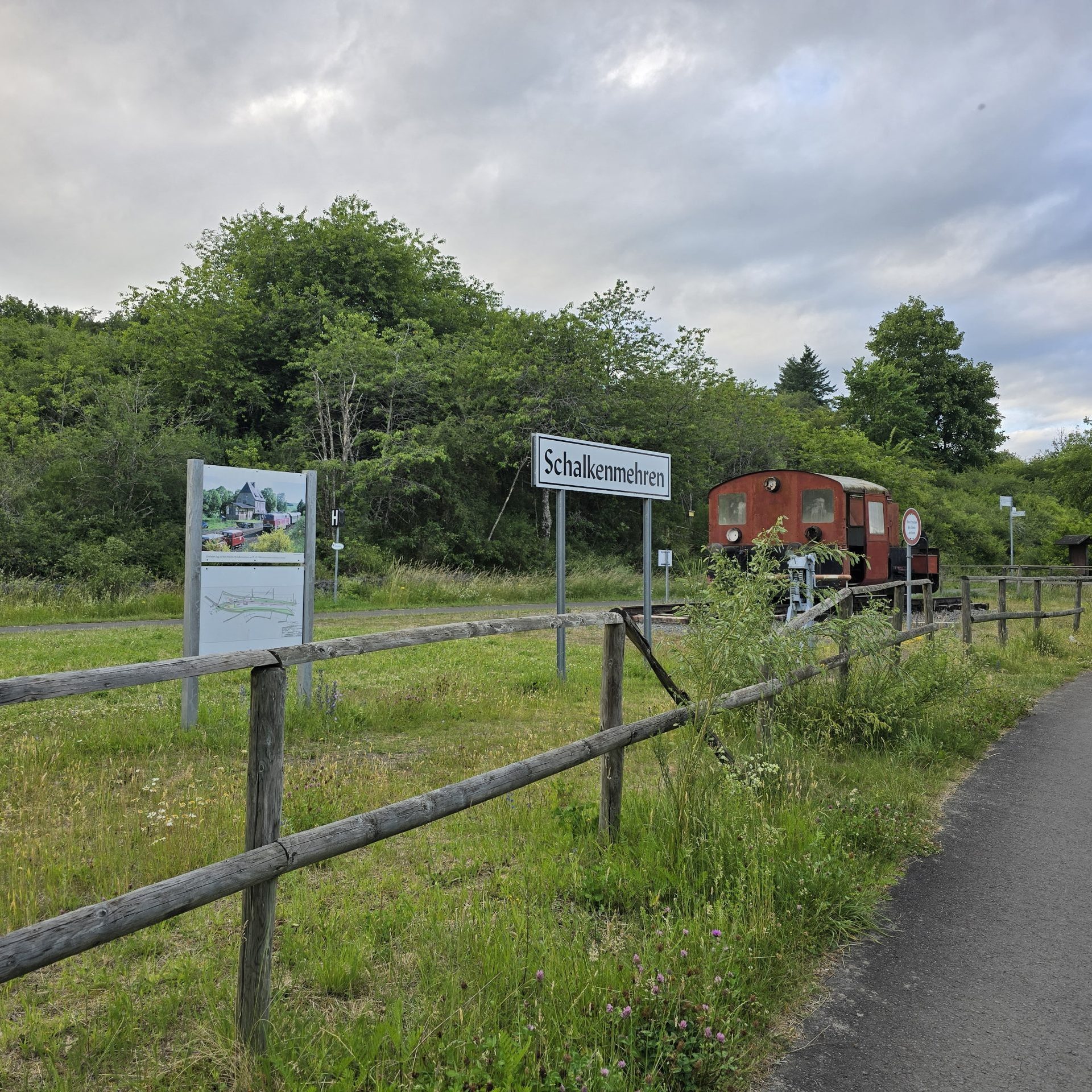 Schilder und alte, verrostete Lok auf einer Wiese, im Vordergrund ein Weg, im Hintergrund Büsche und Bäume