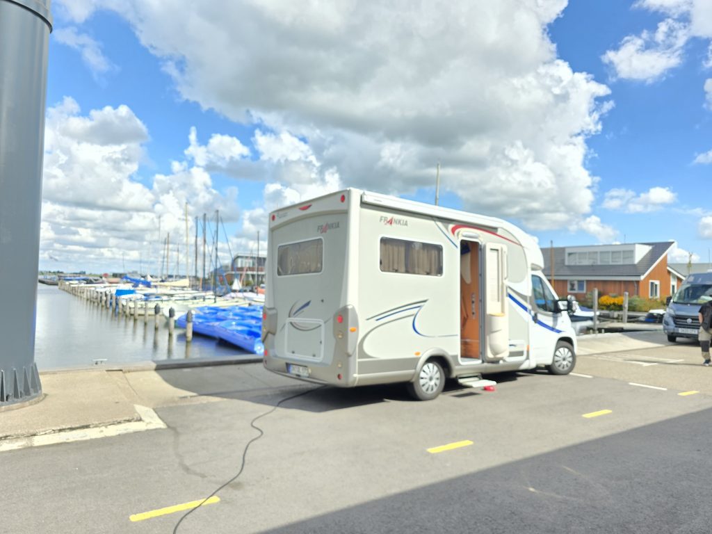 Wohnmobil auf einem Kai direkt am Rand neben dem Wasser und einigen Yachten, darüber blauer Himmer mit weißen Wolken